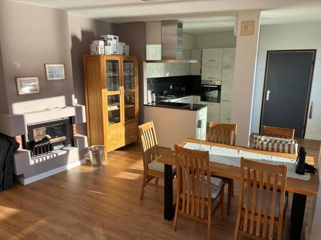 Wohnzimmer mit Kamin, Essbereich und offener Küche; Holzmöbel, neutrale Wände, Sonnenlicht.