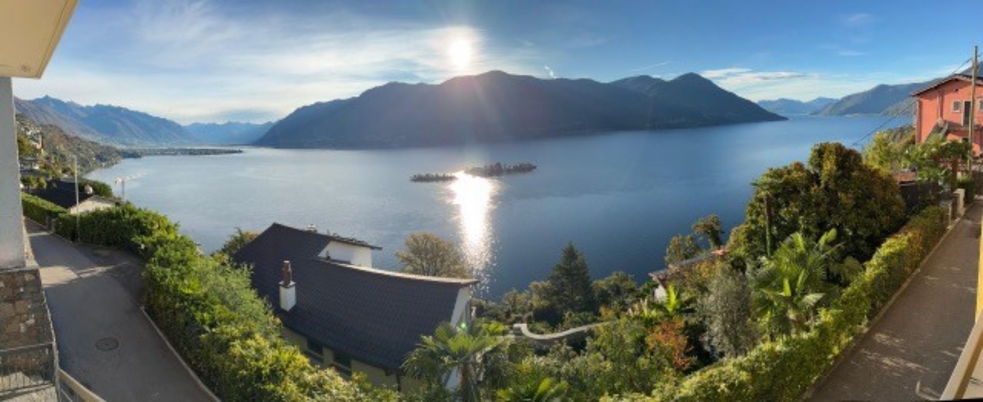 Ein sonniger Seeblick, eingerahmt von grünen Bäumen, mit Bergen im Hintergrund und einem Haus auf der rechten Seite.
