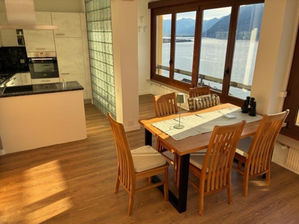 Küche und Essbereich mit Holztisch, Stühlen und grossem Fenster mit Blick auf einen See.