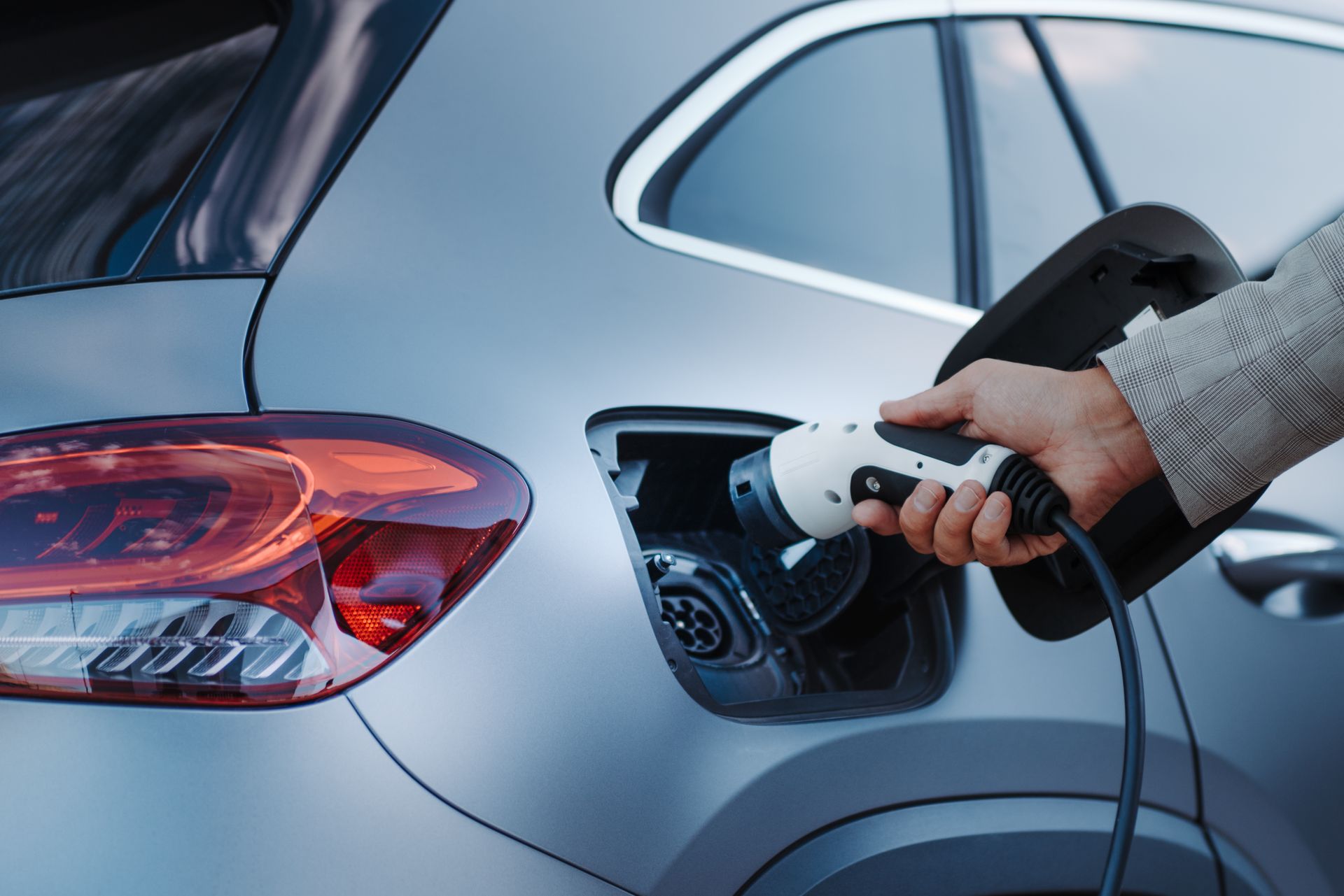 Une personne recharge une voiture électrique argentée avec une prise au niveau du port de recharge arrière