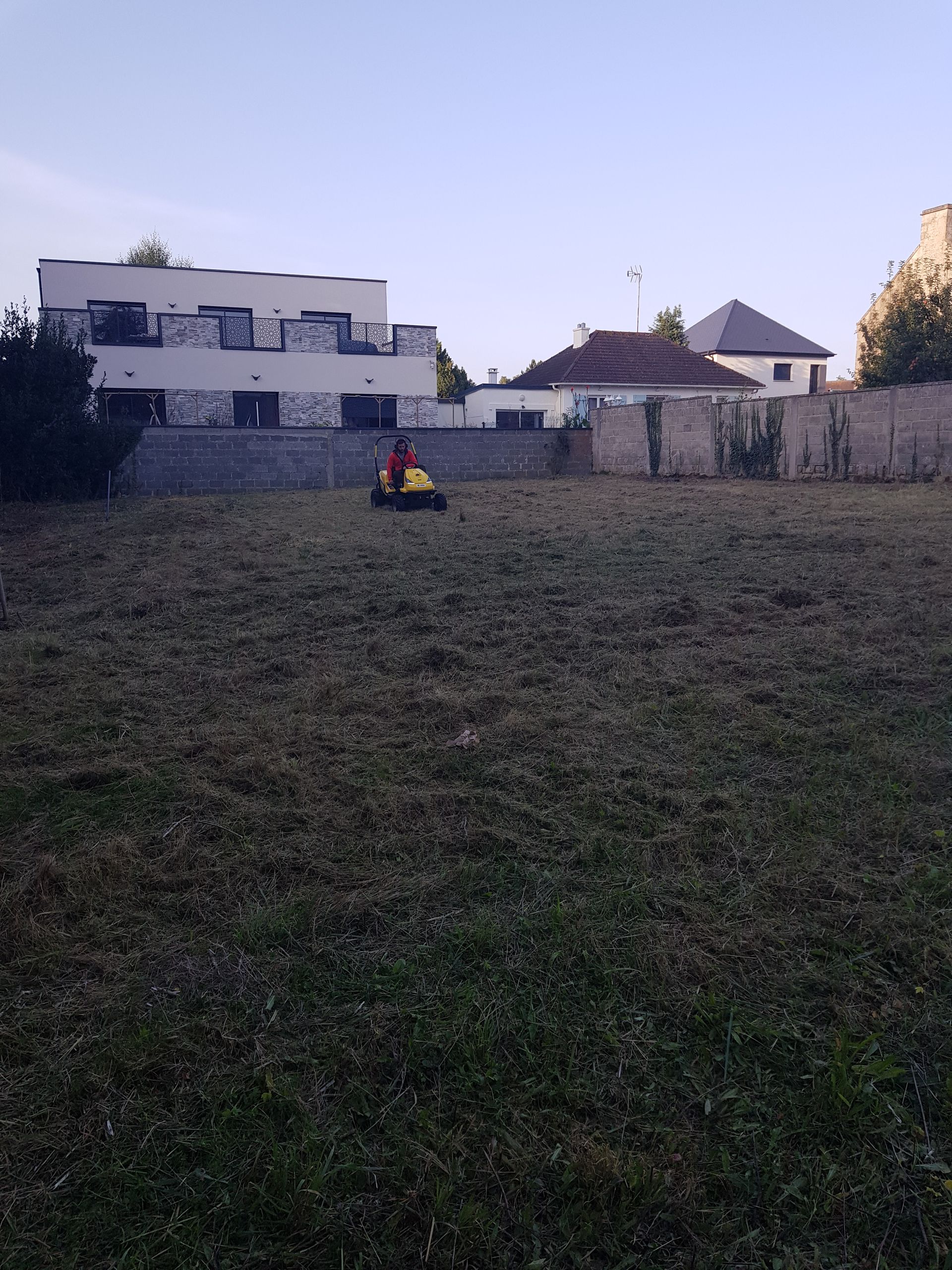 Un enfant conduit un petit véhicule jaune dans une cour herbeuse près de maisons blanches au crépuscule