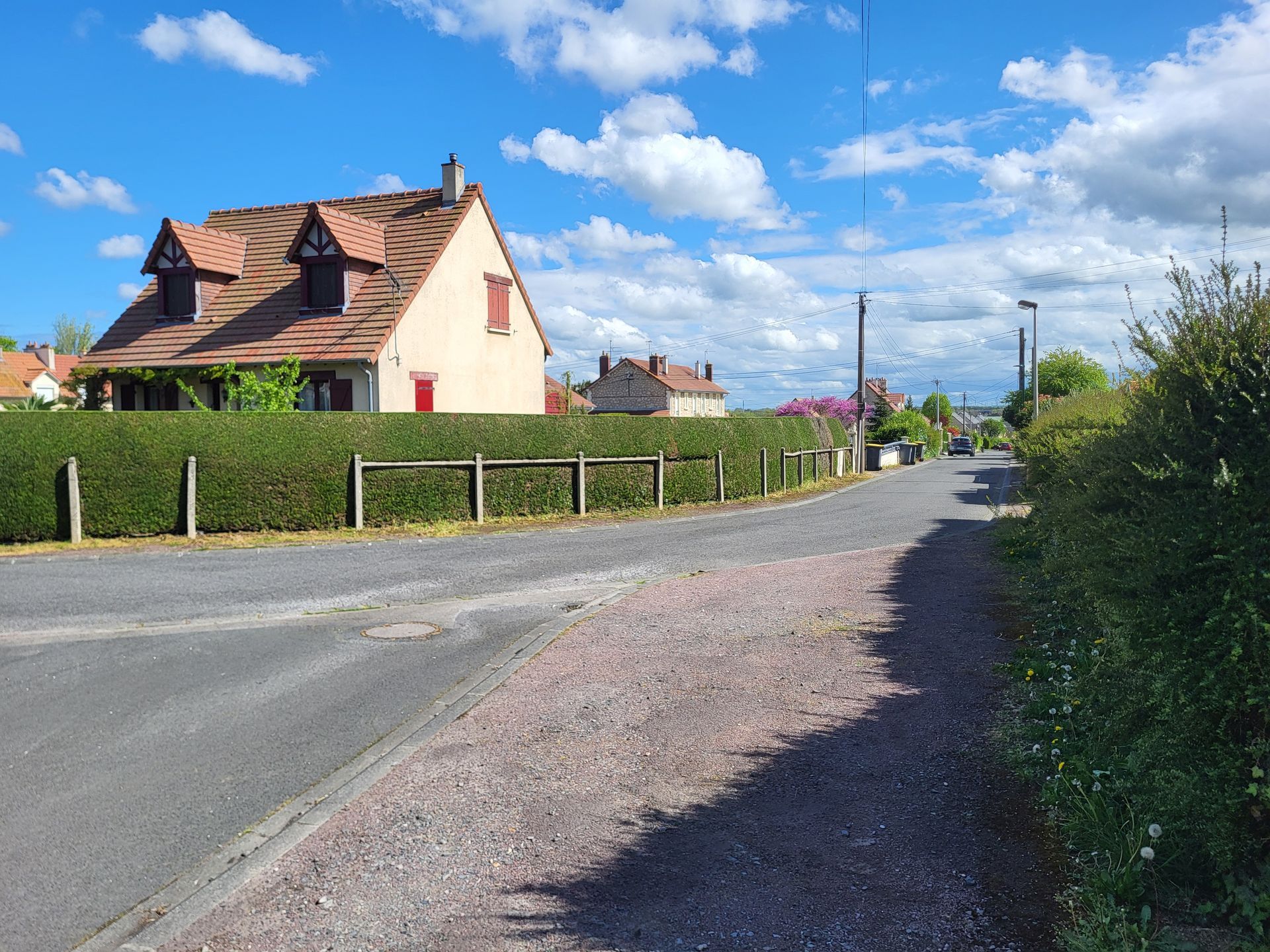 Rue résidentielle tranquille avec une maison couleur crème, des haies et un ciel bleu parsemé de nuages ​​épars.