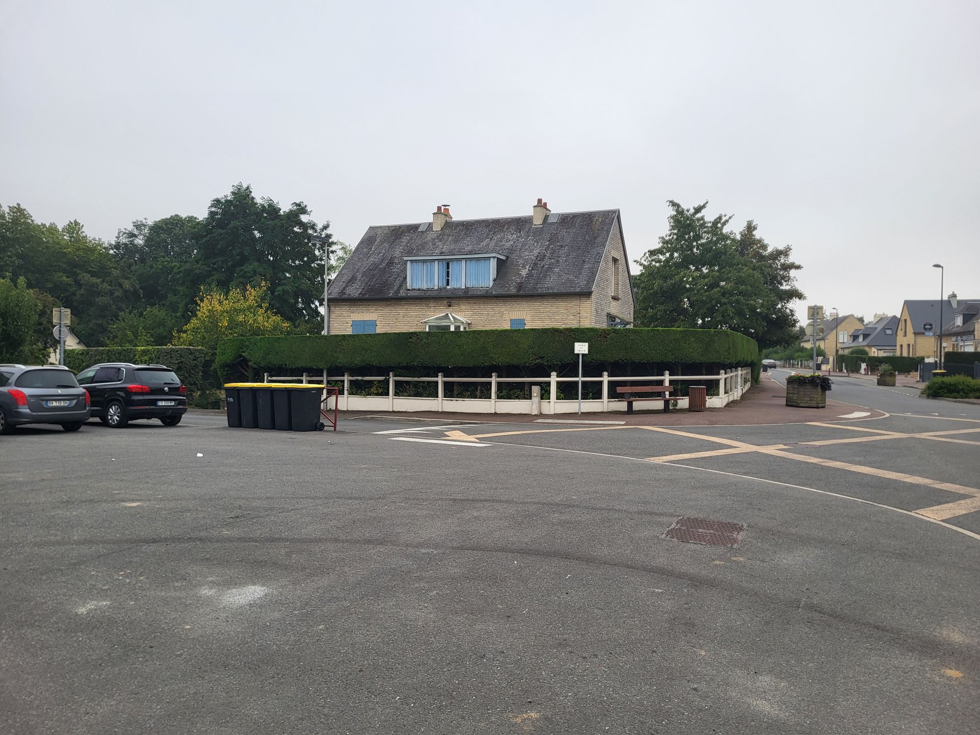 Un parking rural tranquille avec une maison en pierre et quelques voitures sous un ciel couvert.