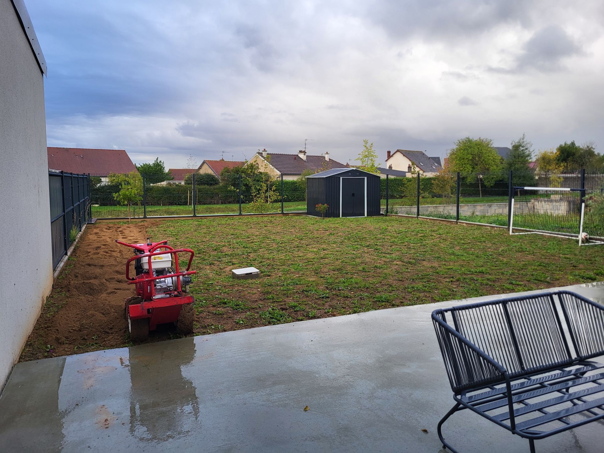 Terrasse arrière avec un barbecue rouge, béton mouillé, pelouse et ciel nuageux.