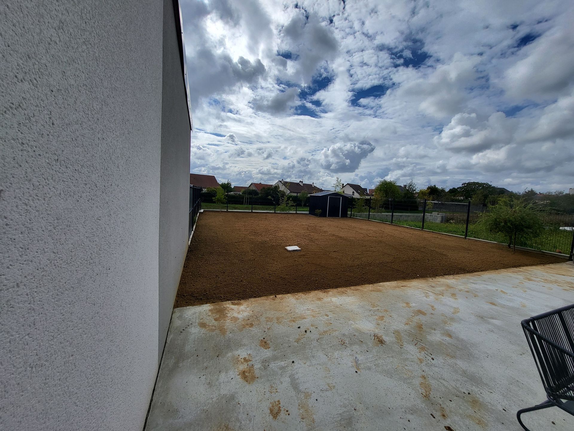 Vaste cour avec terre nue, terrasse en béton et ciel nuageux à côté d'un mur blanc