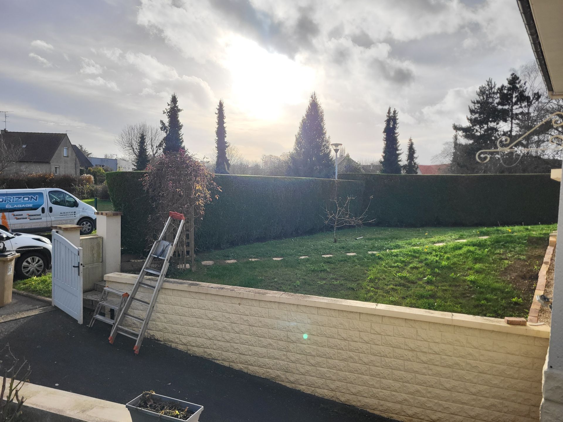 Jardin avec pelouse surélevée derrière un mur de soutènement blanc, une haie, une échelle et un camion sur la gauche sous un ciel nuageux