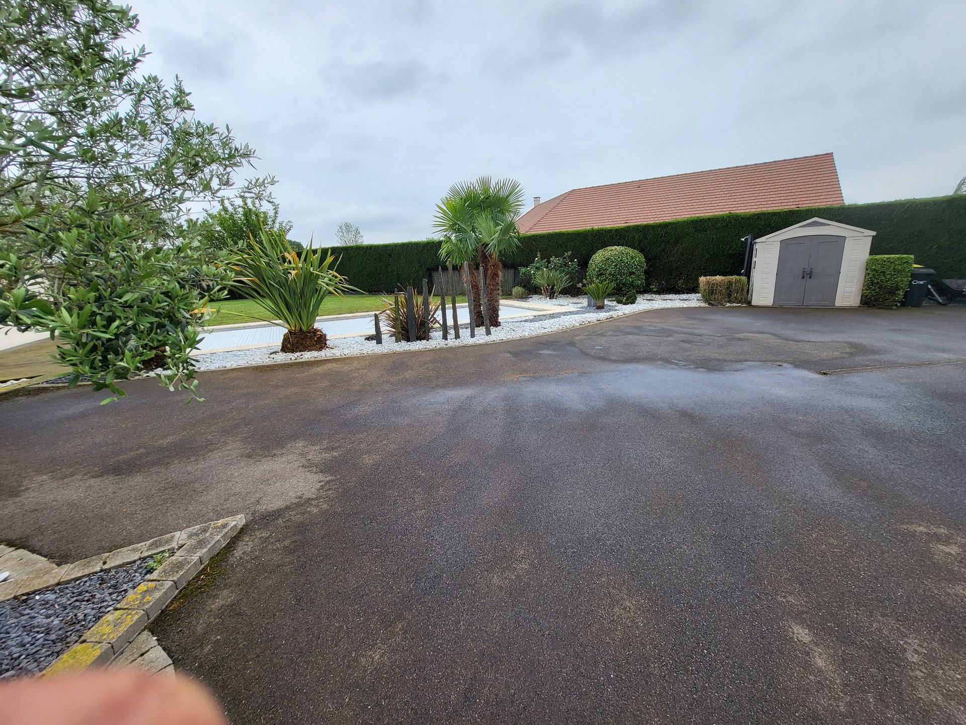 Allée mouillée longeant un jardin paysager avec piscine, arbustes et petit abri de jardin sous un ciel couvert.