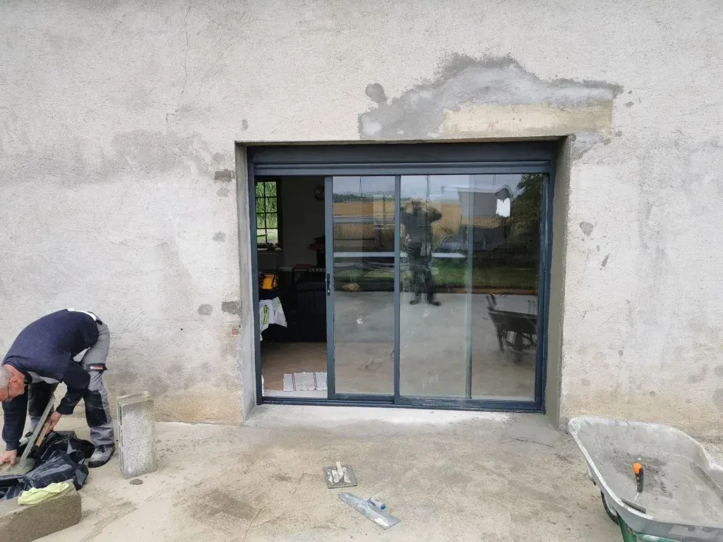 Vue extérieure d'une porte coulissante en verre avec volet ; une personne travaille à proximité, en extérieur.