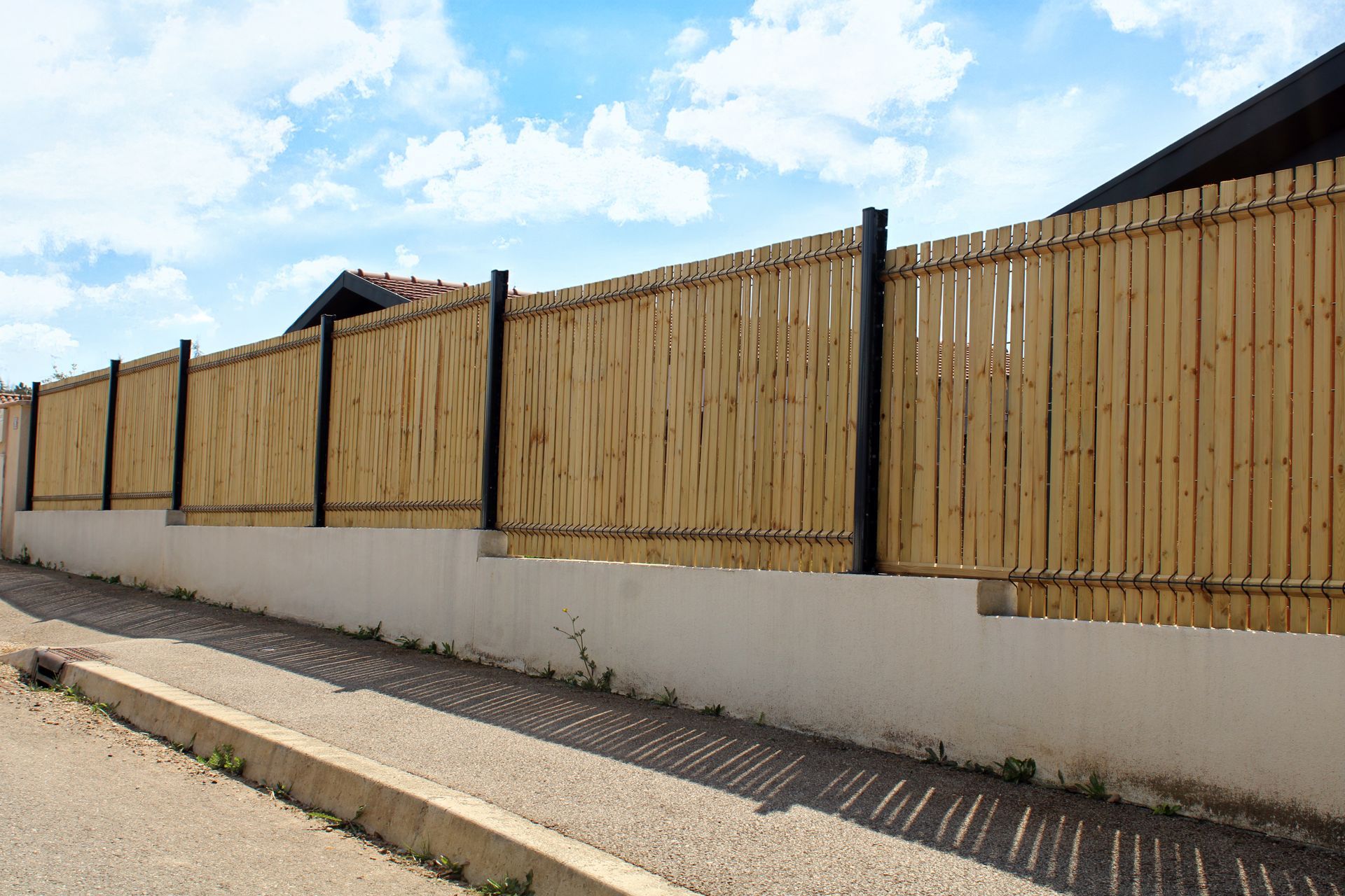 Une clôture en bois surplombe un mur de soubassement blanc, le long d'un trottoir sous un ciel bleu.