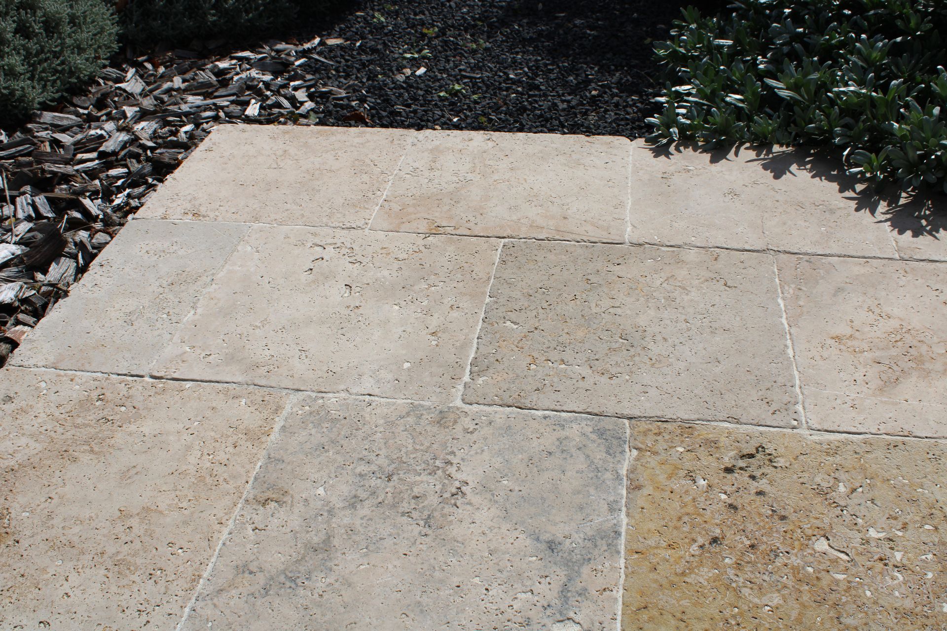 Terrasse en pierre beige avec paillis et galets foncés bordés de feuillage vert.