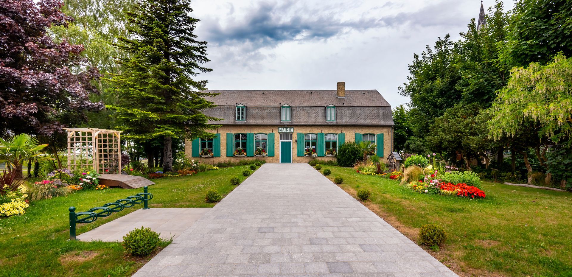 Un long chemin pavé mène à une maison aux volets et à la porte verts, encadrée d'arbres et de fleurs.