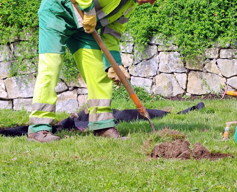 Un ouvrier en tenue de sécurité creuse dans une zone herbeuse avec une pelle.