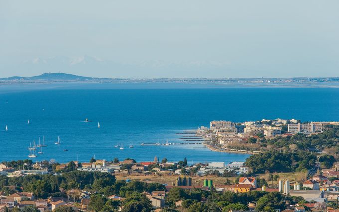 Ville côtière aux eaux bleues et aux voiliers, avec des bâtiments et une montagne à l'horizon.