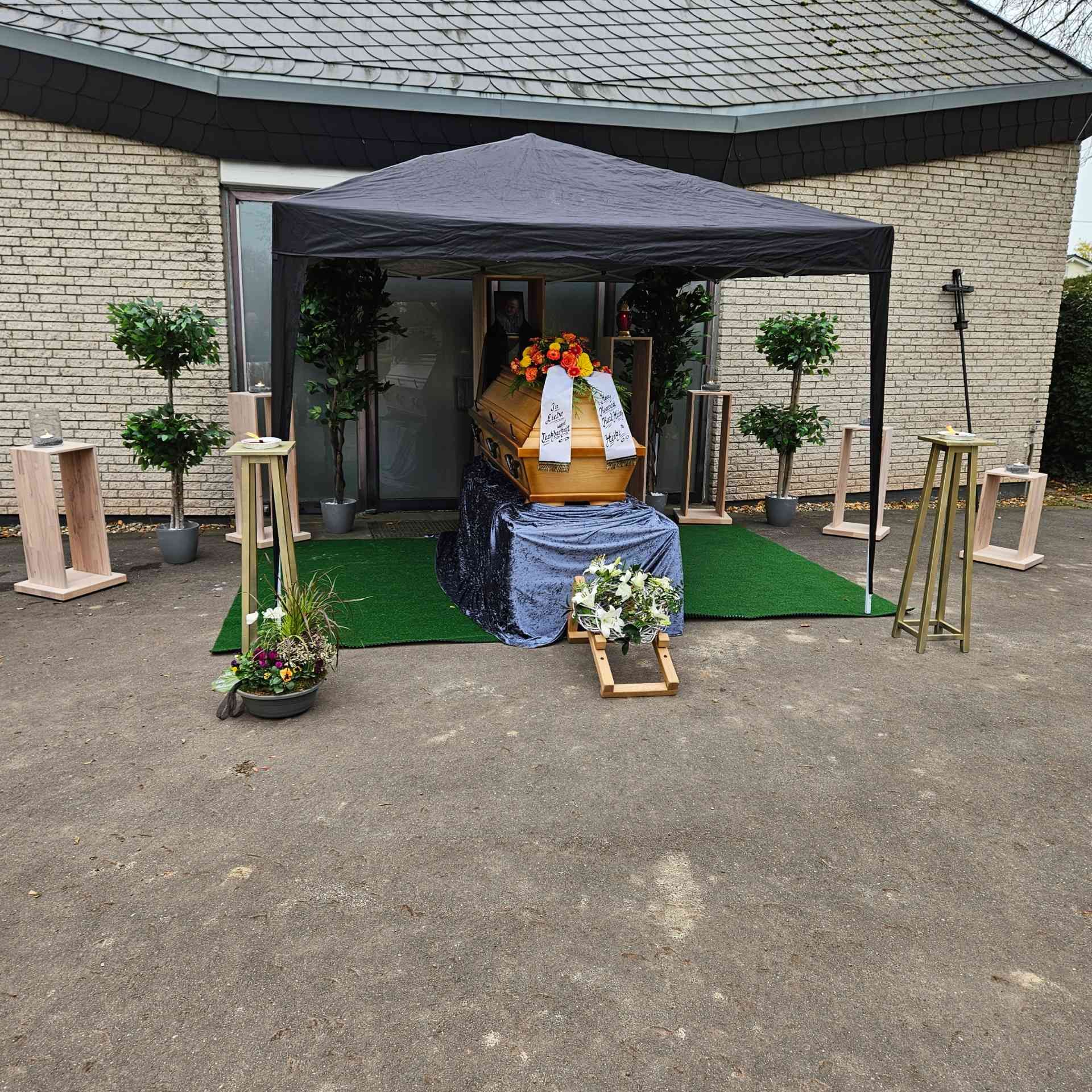 Sarg unter einem schwarzen Zelt vor einem Gebäude, mit Blumen geschmückt, für eine Beerdigung.