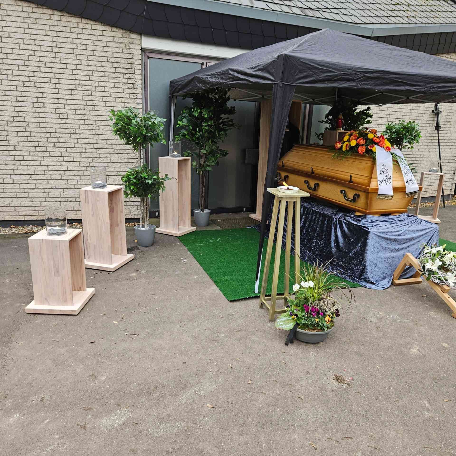 Trauerfeier mit einem Sarg unter einem Baldachin, Blumenarrangements und Holzsockeln vor einem Gebäude.