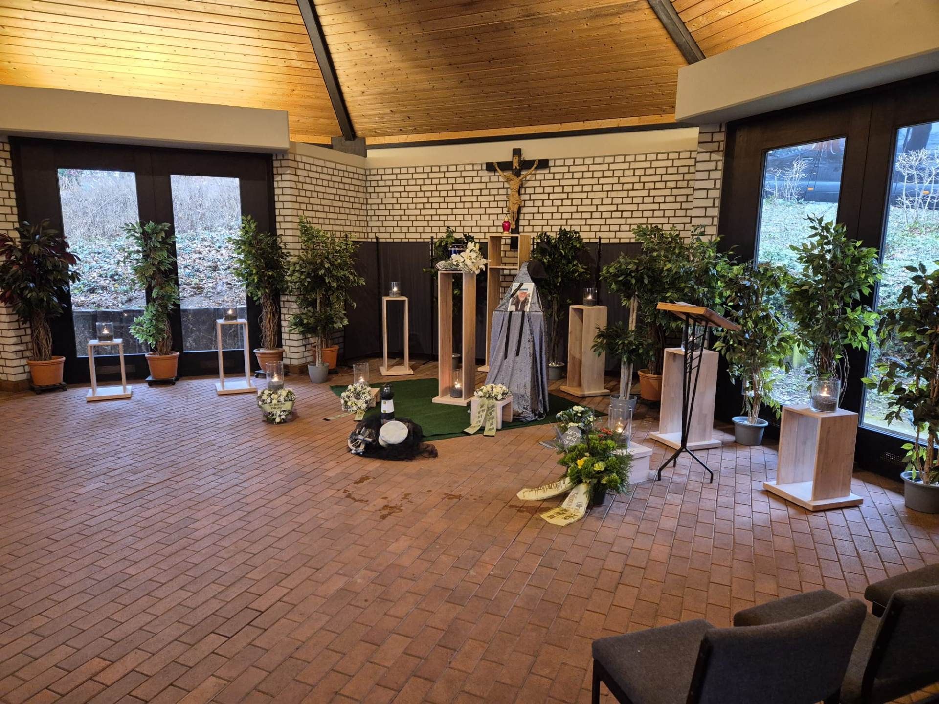 Trauerkapelle mit Ziegelboden, Kreuz, Blumen und Sitzgelegenheiten.
