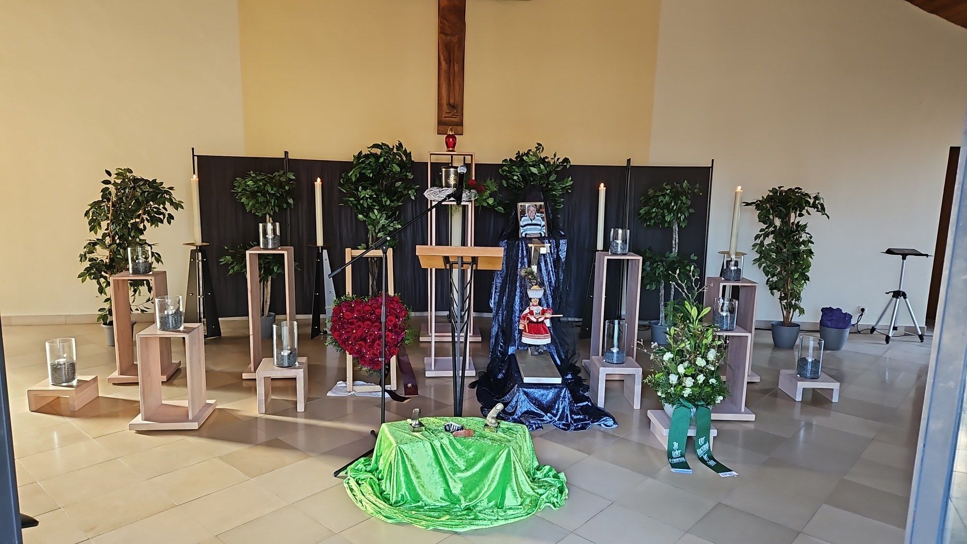 Altar mit Blumenarrangements, Kerzen und einem Kreuz. Grüne Tischdecke davor. Holzsockel.