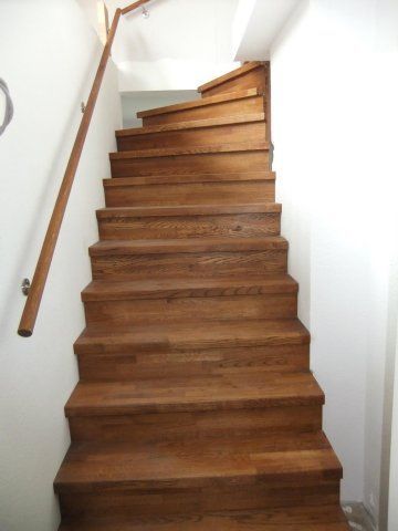 Holztreppe mit Handlauf, die zwischen weißen Wänden nach oben führt.