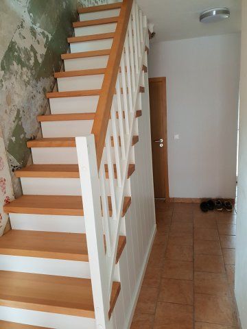 Holztreppe mit weißen Setzstufen, Holzstufen und weißem Geländer. Links beschädigte Wand.