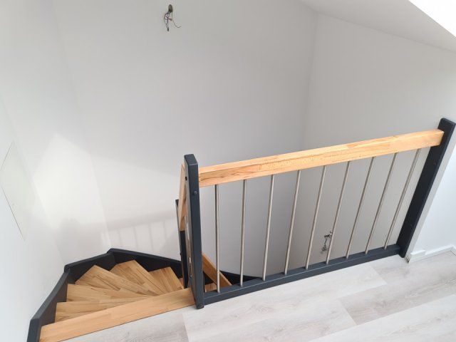 Holztreppe mit schwarzem Geländer, Holzhandlauf und Metallstangen. Weiße Wände.