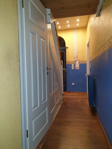 Schmaler Flur mit Holzboden, weißer Tür und gelben und blauen Wänden.