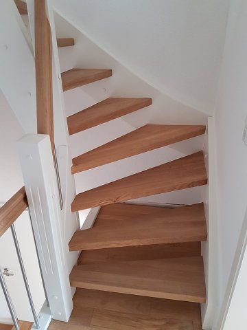 Holztreppe mit abgewinkelten Stufen in einem Raum mit weißen Wänden.