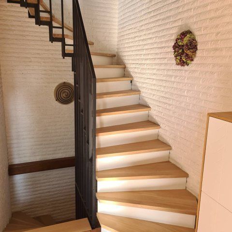 Holztreppe mit weißen Setzstufen und schwarzem Metallgeländer vor einer weißen Ziegelwand.