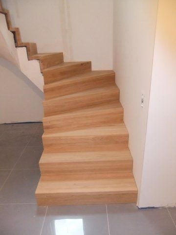 Holztreppe mit geschwungener Kante, helles Holz, vor weißen Wänden, auf gefliestem Boden.