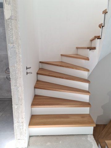 Holztreppe mit weißen Setzstufen vor einer weißen Wand. Die Stufen führen nach oben und sind gewunden.