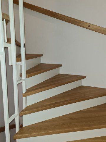 Holztreppe mit weißen Setzstufen und Handlauf vor einer weißen Wand; das rechte Geländer ist aus Holz.