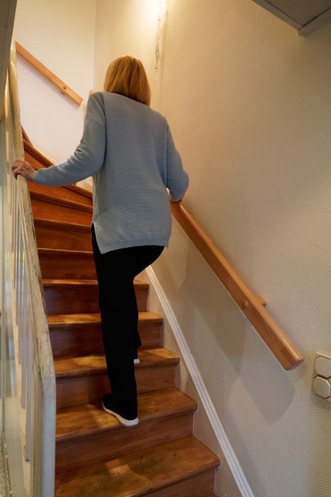 Frau in schwarzen Hosen steigt Holztreppen hinauf und benutzt dabei Handläufe.