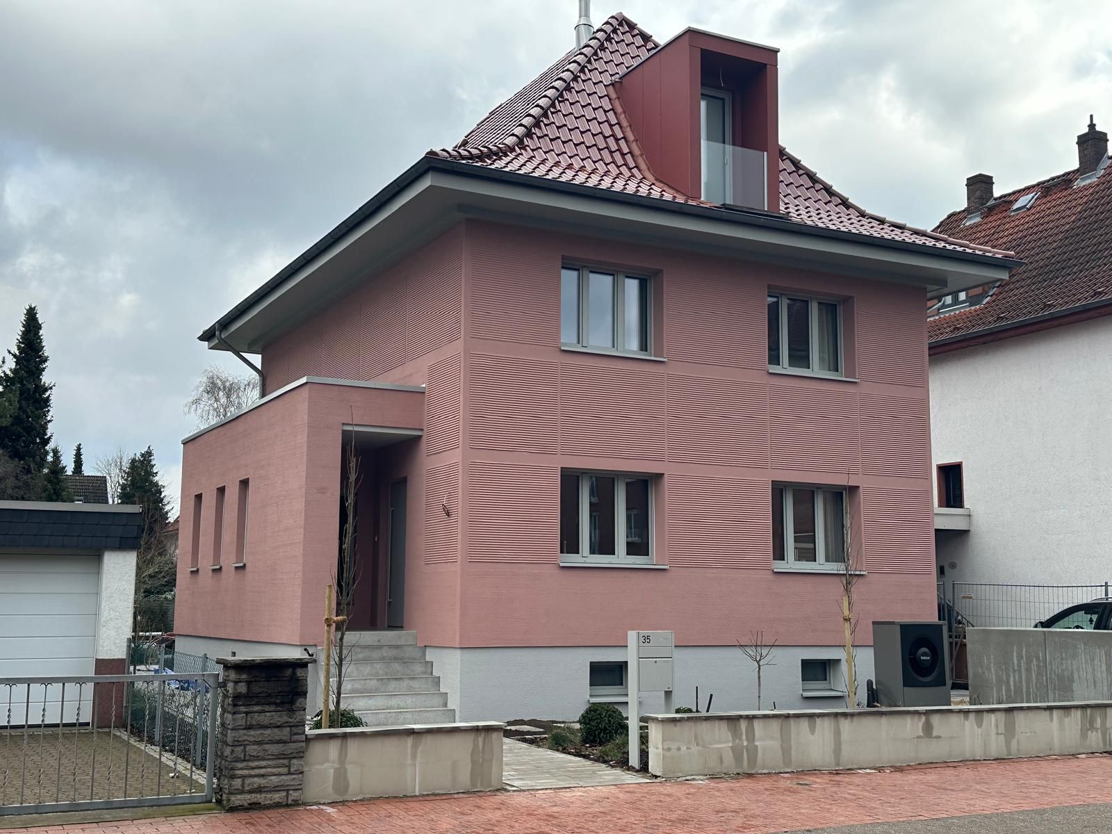 Zweistöckiges rosafarbenes Haus mit rotem Dach, kleinem Vorgarten und grauen Zierelementen unter bewölktem Himmel.