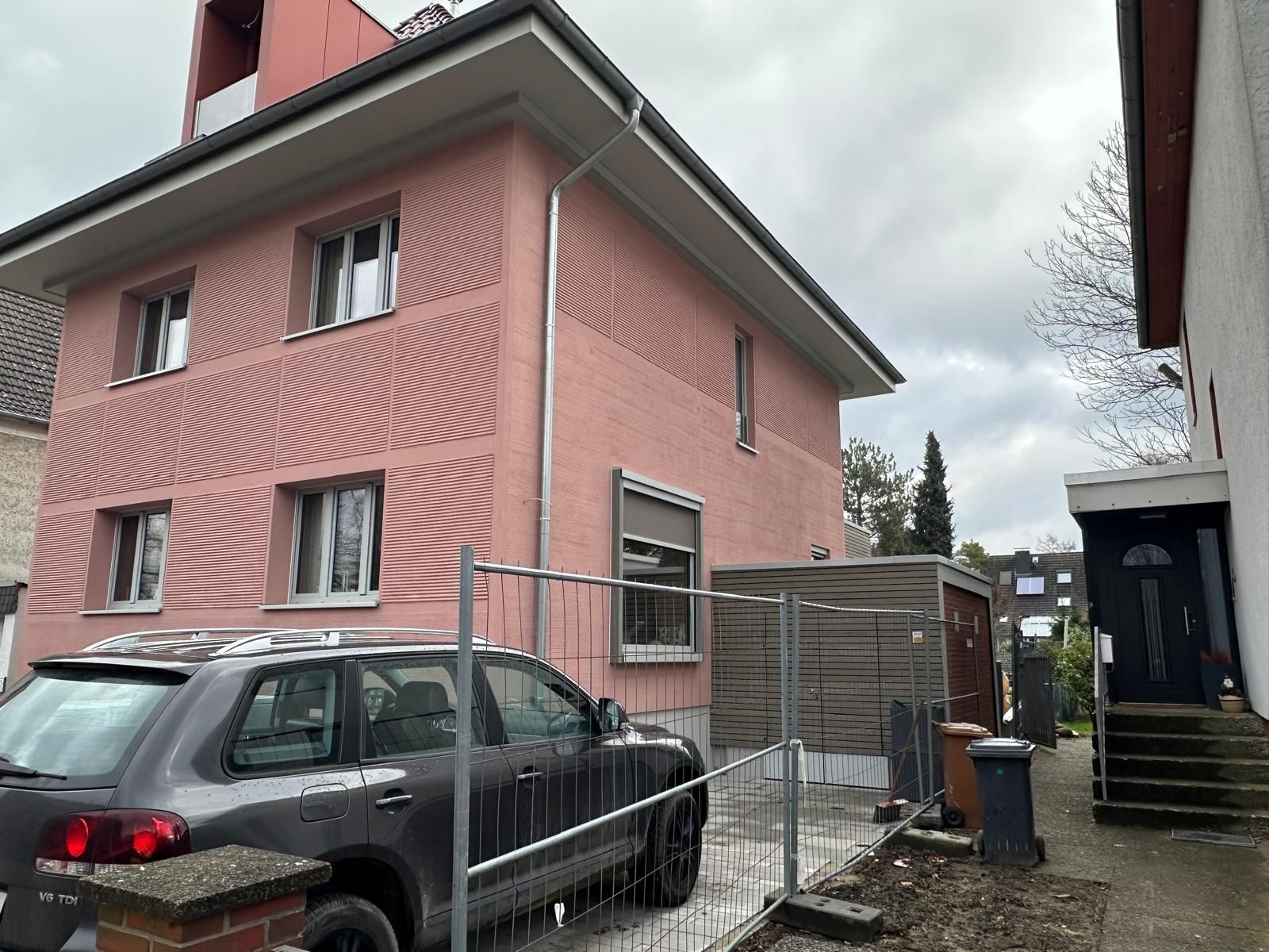 Rosa Gebäude mit einem davor geparkten Auto; grauer Himmel und schmale Gasse zur Rechten.