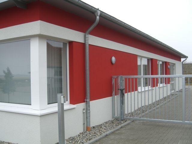 Ein rot-weißes Gebäude mit Fenstern, einem Metalltor und einem grauen Dach vor einem bewölkten Himmel.