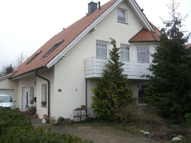 Weißes Haus mit rotem Dach, Balkon und angebauter Garage. Bäume und Auffahrt davor.