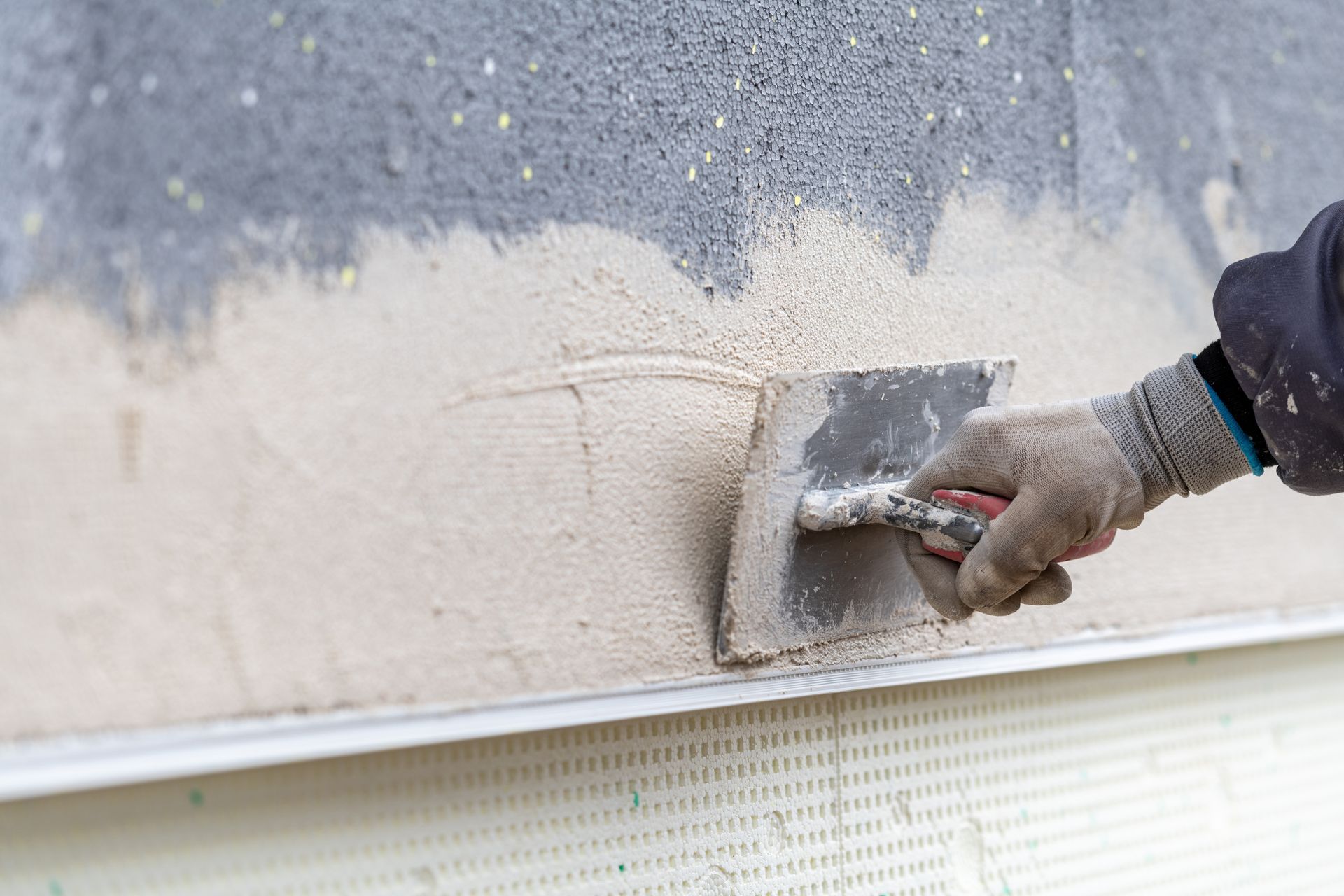Eine Person mit Handschuhen trägt mit einer Kelle hellen Putz auf eine graue Außenwand auf.