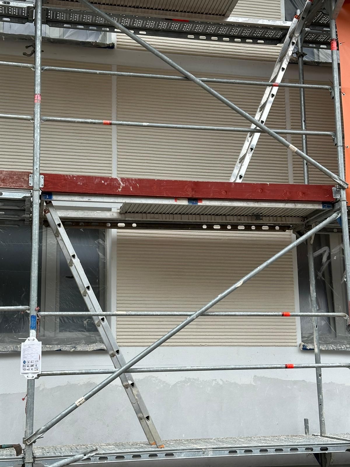 Ein Baugerüst lehnt an einem Gebäude mit geschlossenen Jalousien und einem roten Lichtstrahl.