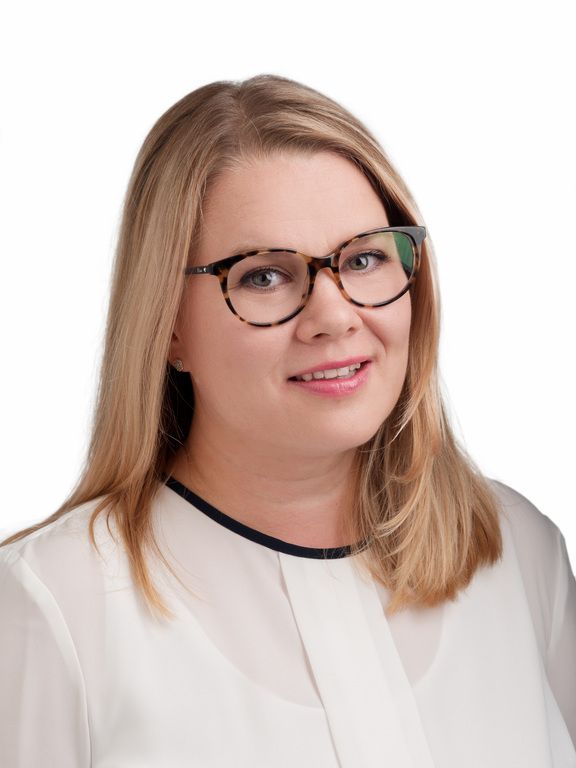 Muotokuva hymyilevästä vaaleatukkaisesta naisesta, jolla on silmälasit ja valkoinen pusero, valkoista taustaa vasten.
