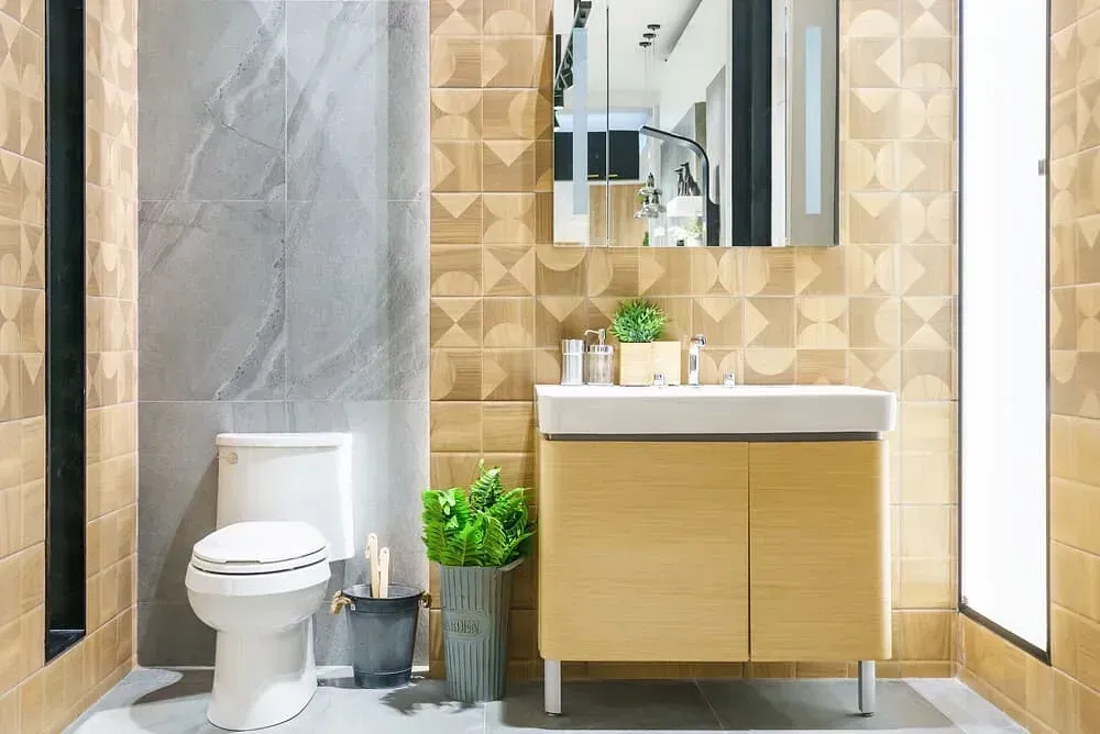 Baño con azulejos dorados y grises, lavabo de madera e inodoro. El espejo refleja el espacio interior.