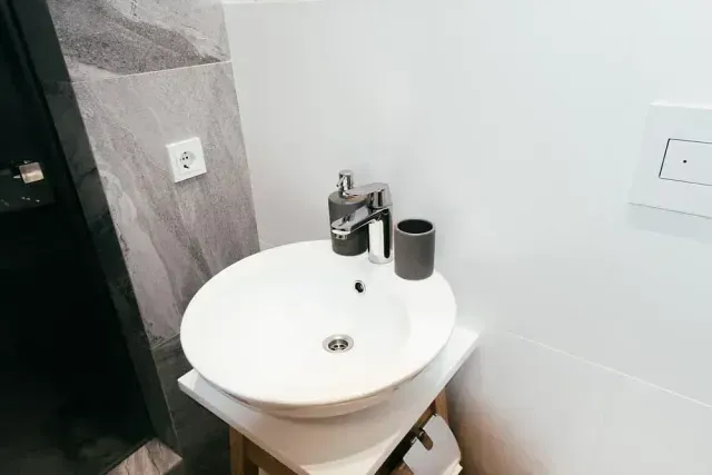 Lavabo redondo blanco sobre un pequeño soporte de madera en un baño. Dispensador de jabón, grifo y vaso sobre la encimera.