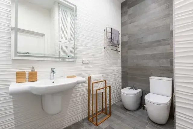 Baño moderno con lavabo e inodoro blancos y paredes texturizadas. Azulejos grises y toallero de madera.