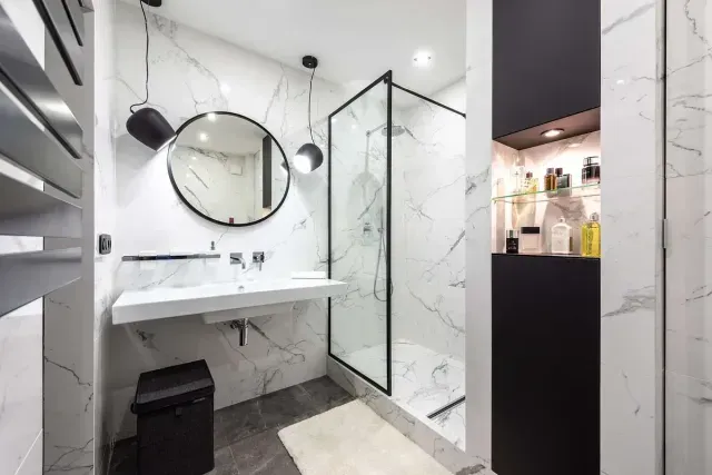 Baño moderno de mármol con detalles en negro, ducha de cristal y espejo redondo.