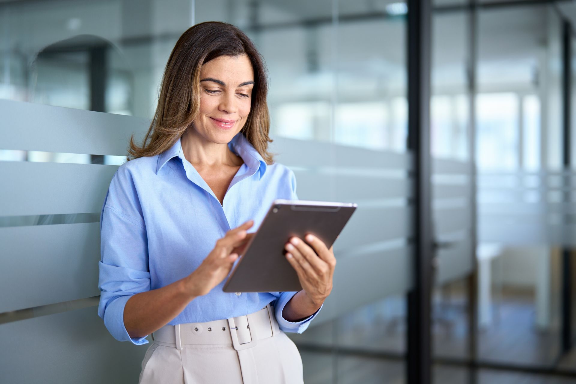 Une femme en chemise bleue et pantalon beige utilise une tablette, souriant près d'une vitre dépolie dans son bureau.