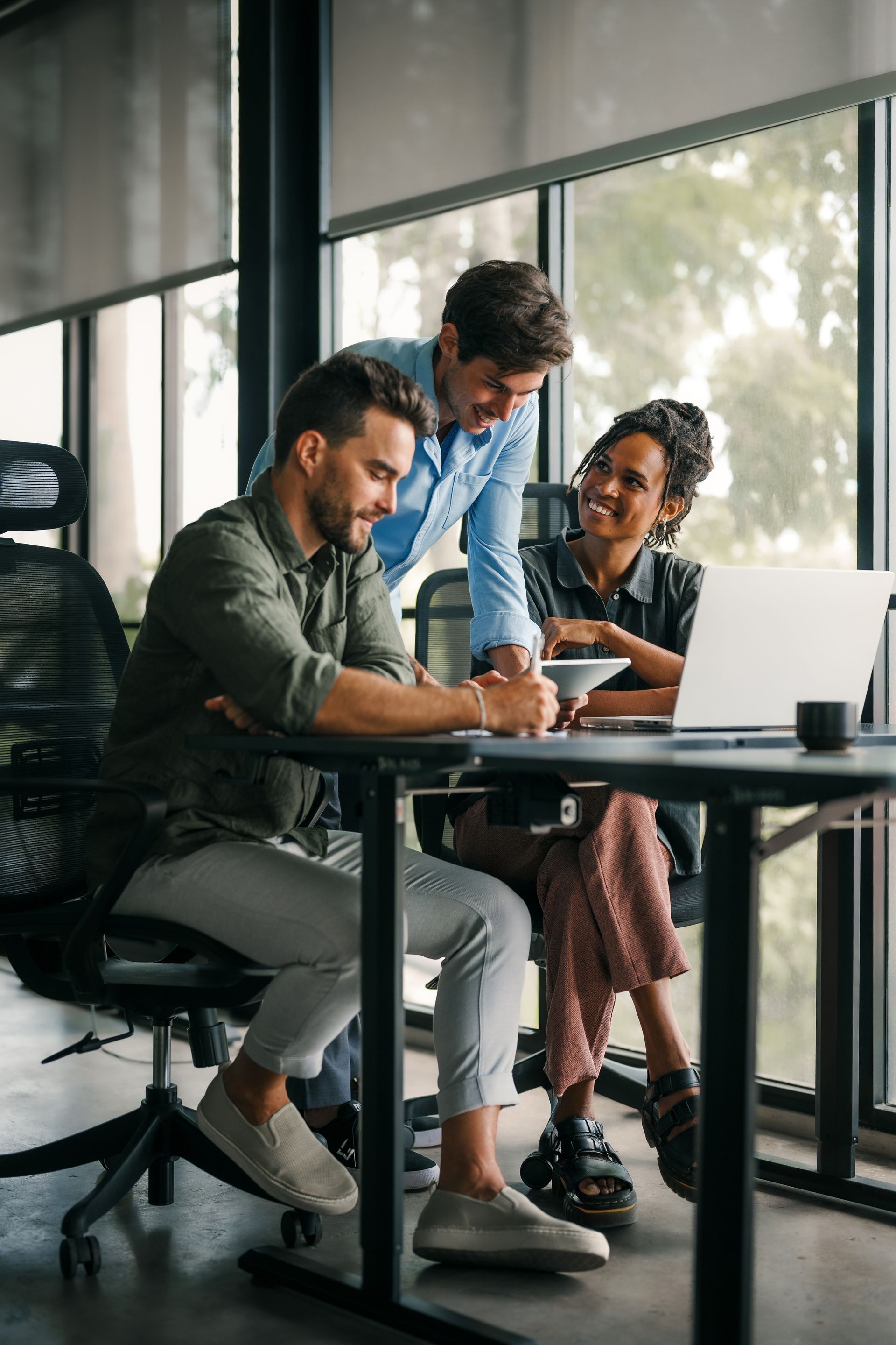 Trois personnes travaillent ensemble à un bureau près d'une fenêtre ; l'une est assise, les deux autres sont debout. Elles se concentrent sur l'écran d'un ordinateur portable et sur des documents.