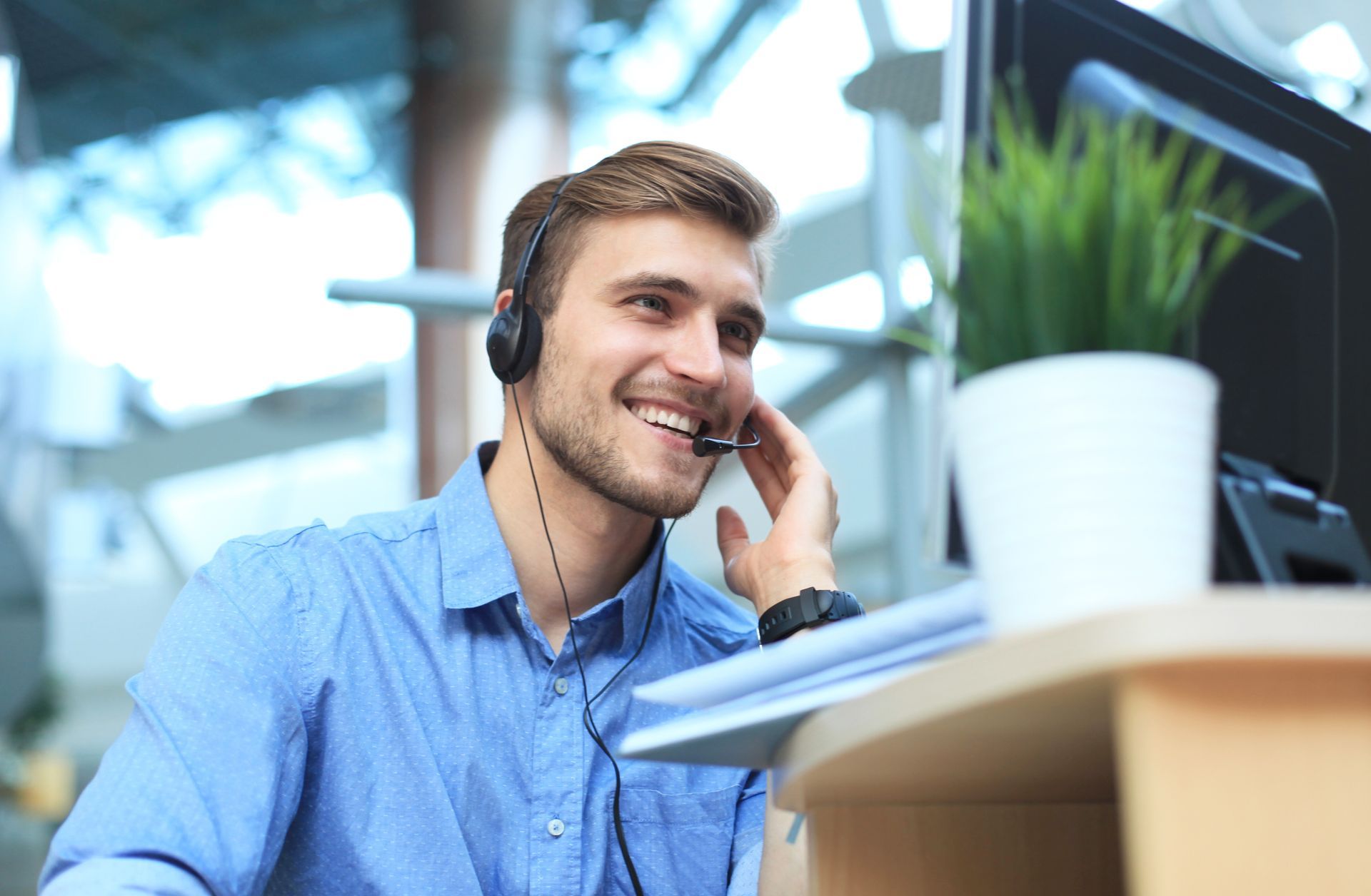 Un homme portant un casque audio sourit, assis à un bureau avec un ordinateur et une plante.