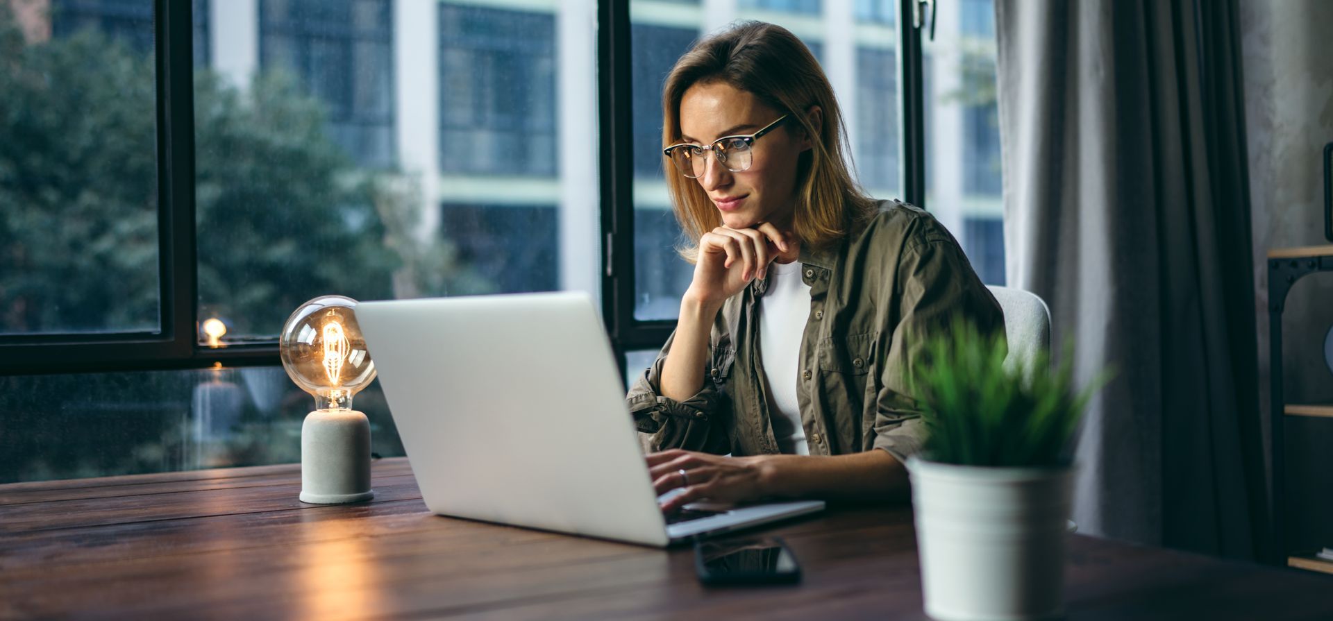 Une femme portant des lunettes travaille sur un ordinateur portable à un bureau près d'une fenêtre. Une lampe et une plante sont posées sur le bureau.