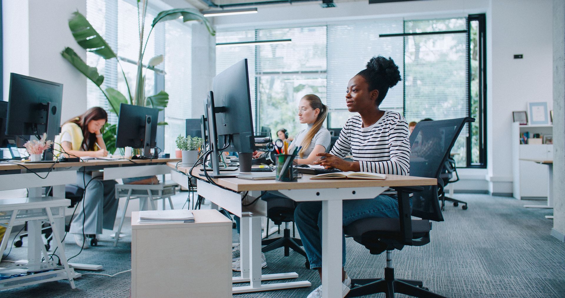 Des personnes travaillent à des bureaux dans un espace de travail lumineux. Une femme regarde et fait des gestes. D'autres utilisent des ordinateurs.