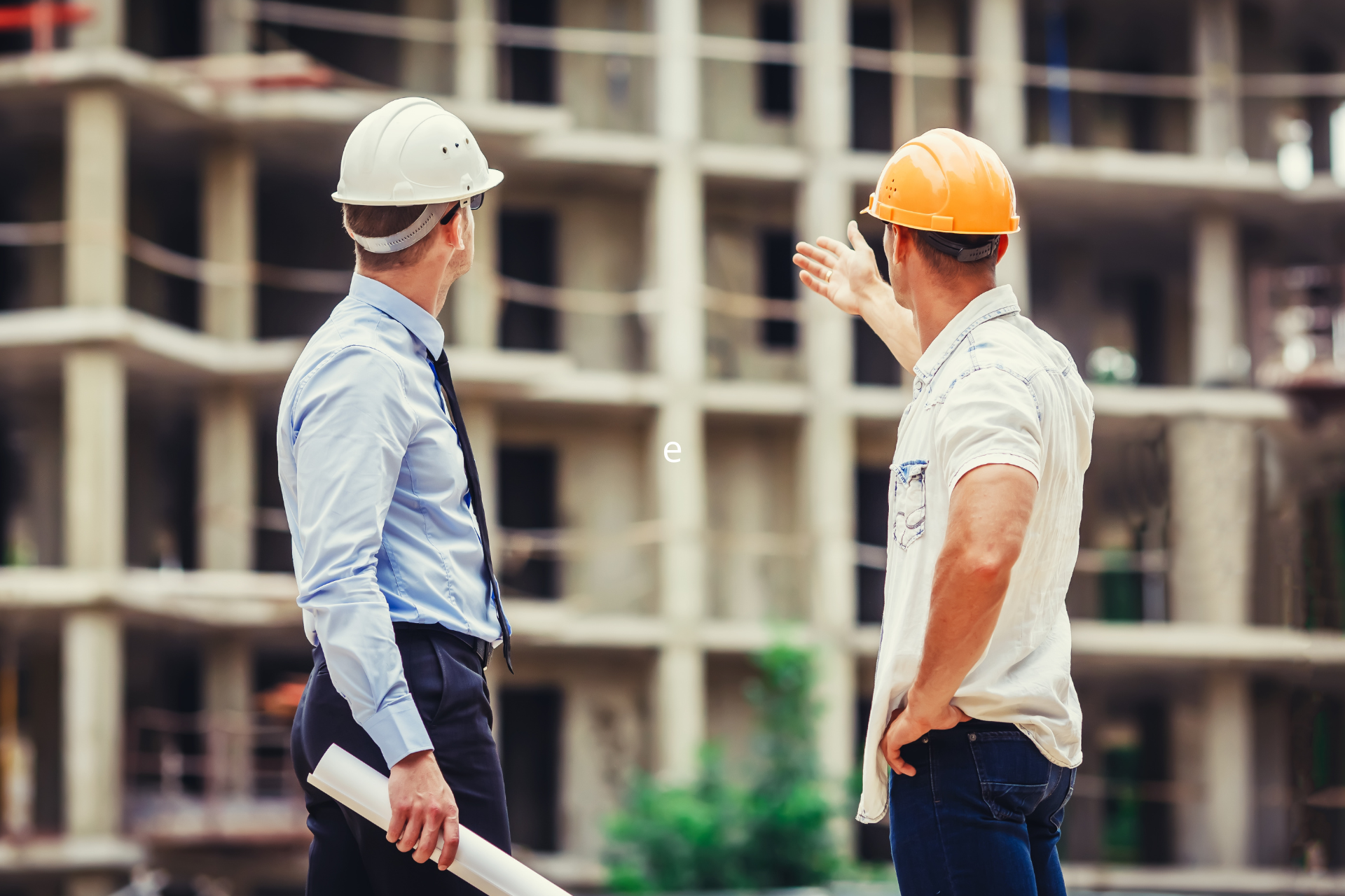 Dos obreros de la construcción con cascos observan un edificio en construcción; uno de ellos señala.