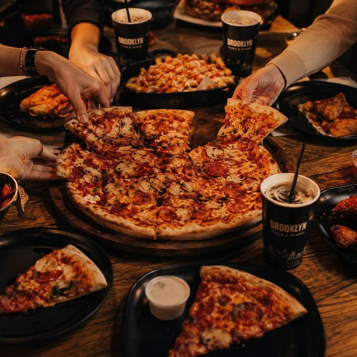 Manos que buscan porciones de pizza de queso en una mesa abarrotada de bebidas.