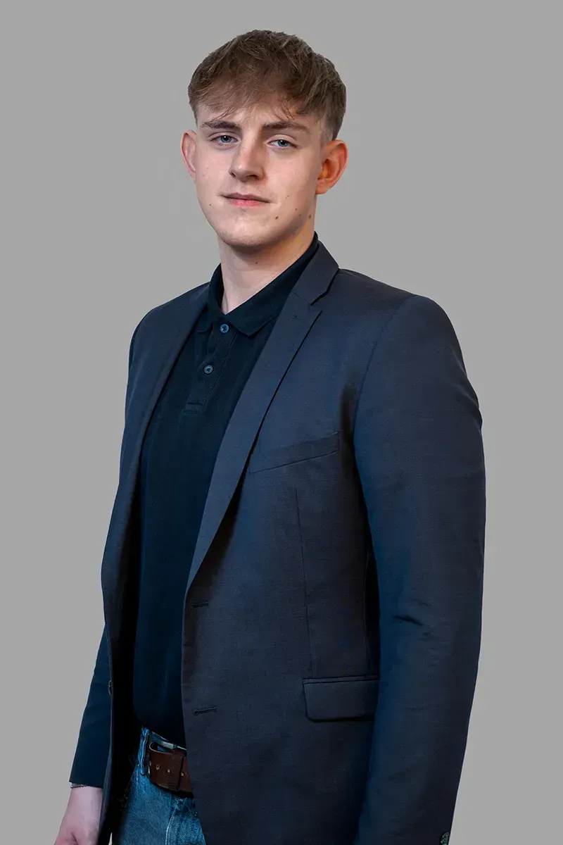 Ein junger Mann in Anzug und schwarzem Hemd steht vor einem grauen Hintergrund.